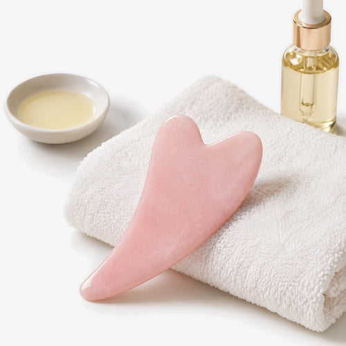 Gua Sha Stone — płytka z różowego kwarcu na ręczniku, obok olejek do twarzy