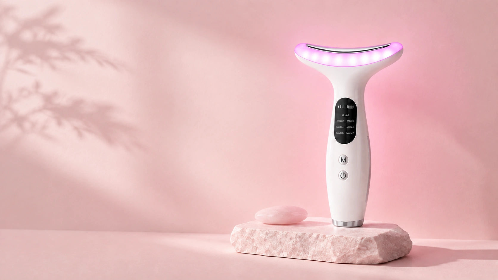LED neck and face massager glowing on a rose quartz podium beside a gua sha stone